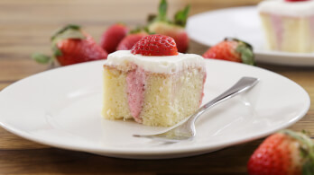 A slice of strawberry shortcake topped with a fresh strawberry on a white plate. The cake has a layer of pink filling and is garnished with white frosting. A fork is placed on the plate, and whole strawberries are scattered in the background.