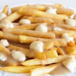 A plate of poutine, featuring golden French fries topped with cheese curds and smothered in light brown gravy, is shown on a white plate with blue decorative trim.