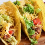 Three crispy tacos filled with ground meat, shredded cheddar cheese, chopped lettuce, and diced tomatoes are displayed on a wooden surface. The tacos are arranged in a line, showcasing their colorful and fresh ingredients.