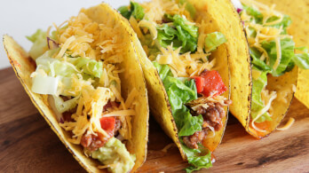 Three crispy tacos filled with ground meat, shredded cheddar cheese, chopped lettuce, and diced tomatoes are displayed on a wooden surface. The tacos are arranged in a line, showcasing their colorful and fresh ingredients.