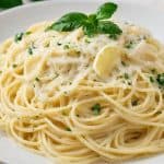 A plate of spaghetti with a creamy sauce, garnished with grated cheese, fresh parsley, and a lemon slice. A sprig of basil is placed on top for decoration. Sliced lemons and parsley leaves are visible in the background.