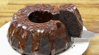 A rich chocolate bundt cake topped with glossy chocolate ganache and chocolate sprinkles sits on a white plate. A slice is being lifted out with a silver cake server, revealing the moist, dark interior. The background is a wooden table.