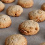 A close-up view of freshly baked, round cookies arranged on a sheet of parchment paper. The cookies are golden brown with a slightly cracked surface, indicating a crispy texture.