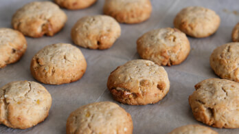 A close-up view of freshly baked, round cookies arranged on a sheet of parchment paper. The cookies are golden brown with a slightly cracked surface, indicating a crispy texture.