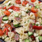 A bowl of pasta salad featuring bowtie pasta, cherry tomatoes, cucumber slices, cubes of mozzarella cheese, and slices of salami, garnished with chopped herbs and placed on a wooden surface.