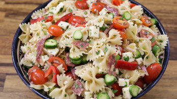 A bowl of pasta salad featuring bowtie pasta, cherry tomatoes, cucumber slices, cubes of mozzarella cheese, and slices of salami, garnished with chopped herbs and placed on a wooden surface.