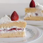 Two slices of sponge cake layered with whipped cream and fresh strawberries are served on white plates. Each slice is topped with a dollop of whipped cream and a whole strawberry. A gold fork is placed beside one of the plates.