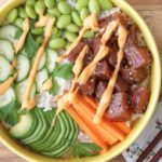 A colorful bowl of poke, featuring cubed pieces of seasoned raw fish, sliced cucumber, edamame, shredded carrots, and white rice, drizzled with a creamy sauce. The bowl is on a wooden surface with floral-patterned napkins and a pair of wooden chopsticks beside it.