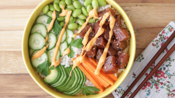 A colorful bowl of poke, featuring cubed pieces of seasoned raw fish, sliced cucumber, edamame, shredded carrots, and white rice, drizzled with a creamy sauce. The bowl is on a wooden surface with floral-patterned napkins and a pair of wooden chopsticks beside it.