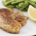 A plate with a piece of cooked fish, a wedge of lemon, and a serving of green beans. The fish appears to be pan-fried with a golden-brown crust. The lemon wedge and green beans are placed to the side.