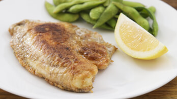 A plate with a piece of cooked fish, a wedge of lemon, and a serving of green beans. The fish appears to be pan-fried with a golden-brown crust. The lemon wedge and green beans are placed to the side.