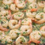 A pan filled with cooked shrimp in a garlic butter sauce, garnished with chopped parsley. The shrimp are evenly distributed and have a light pink color, indicating they are fully cooked. The sauce appears creamy and is sprinkled with herbs and red pepper flakes.