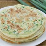 A white plate holds a stack of Chinese scallion pancakes with visible green onion bits. The pancakes are on a wooden table, and several whole green onions are placed next to the plate.