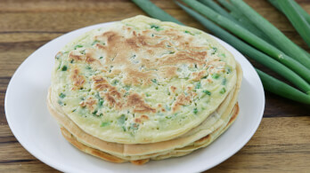 A white plate holds a stack of Chinese scallion pancakes with visible green onion bits. The pancakes are on a wooden table, and several whole green onions are placed next to the plate.