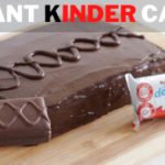 A giant chocolate cake sits on a wooden board, covered in glossy chocolate icing and decorated with a drizzled pattern. In the foreground, a hand holds a smaller, pre-packaged chocolate treat named "Kinder Delice." The text above reads "GIANT KINDER CAKE.