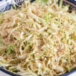 A bowl filled with a cabbage salad topped with sesame seeds. The shredded cabbage is mixed with a dressing, and the salad appears fresh and vibrant. The bowl is blue with a decorative pattern on the rim.