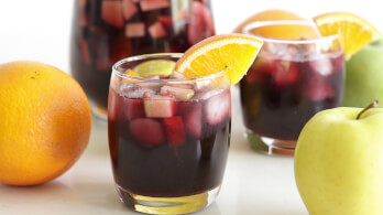 A close-up of two glasses filled with sangria, garnished with orange slices, and containing ice cubes and fruit chunks. One green apple and one orange are on the left, and another fruit (partially visible) is on the right. A pitcher with sangria is in the background.