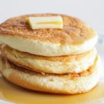 A stack of three fluffy pancakes topped with a square of melting butter and drizzled with a generous amount of syrup, served on a white plate with a subtle blue floral pattern around the rim.