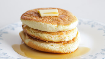 A stack of three fluffy pancakes topped with a square of melting butter and drizzled with a generous amount of syrup, served on a white plate with a subtle blue floral pattern around the rim.