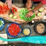 Two people prepare a variety of fresh vegetables on a wooden surface. Ingredients include chopped red peppers, avocado, lettuce, cucumbers, cherry tomatoes, and a pan with seeds. Various bowls and a large wooden paddle are also visible in the scene.