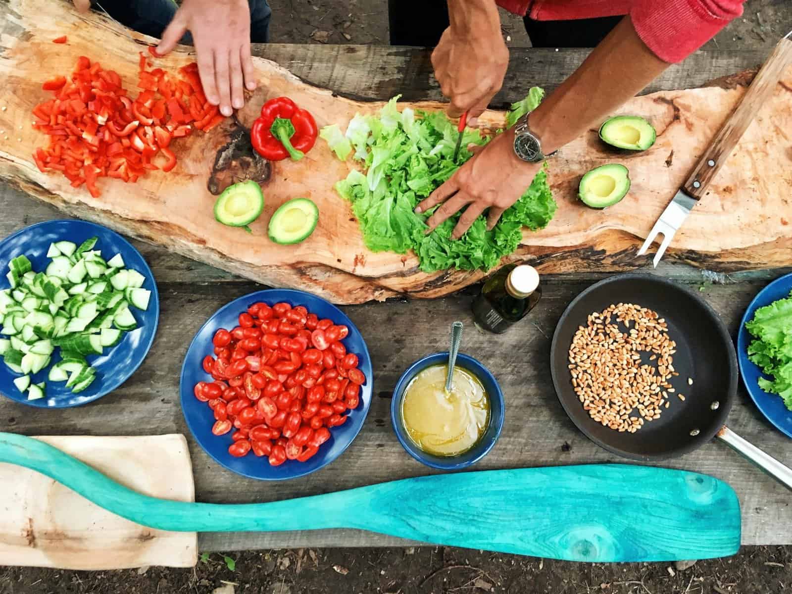 Two people prepare a variety of fresh vegetables on a wooden surface. Ingredients include chopped red peppers, avocado, lettuce, cucumbers, cherry tomatoes, and a pan with seeds. Various bowls and a large wooden paddle are also visible in the scene.