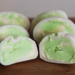 Close-up of multiple pieces of green tea mochi ice cream, some cut in half to show the green ice cream filling. The mochi pieces are resting on a wooden surface.