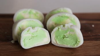 Close-up of multiple pieces of green tea mochi ice cream, some cut in half to show the green ice cream filling. The mochi pieces are resting on a wooden surface.