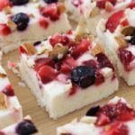 A close-up of several square pieces of frozen yogurt bark lying on a wooden surface. Each piece is topped with a mixture of blueberries, strawberries, and chopped nuts. The bark has a creamy white base.