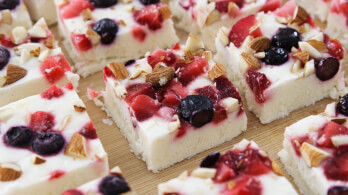 A close-up of several square pieces of frozen yogurt bark lying on a wooden surface. Each piece is topped with a mixture of blueberries, strawberries, and chopped nuts. The bark has a creamy white base.