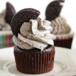 A chocolate cupcake topped with cookies and cream frosting, garnished with half of a chocolate sandwich cookie, sits on a white plate. Other similar cupcakes are slightly blurred in the background, displayed on a light-colored surface.