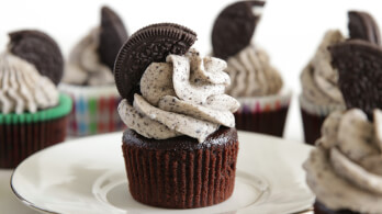 A chocolate cupcake topped with cookies and cream frosting, garnished with half of a chocolate sandwich cookie, sits on a white plate. Other similar cupcakes are slightly blurred in the background, displayed on a light-colored surface.