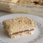 A slice of layered dessert consisting of cream and chocolate layers sits on a white plate with a blue floral border. The dessert has a crumbly top layer. In the background, there is a glass dish containing the remaining dessert. A fork is placed beside the plate.