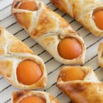 Freshly baked pastries with golden-brown flaky crusts are cooling on a wire rack. Each pastry is filled with a halved apricot, showcasing vibrant orange fruit against the light, crisp dough. The pastries are evenly spaced on the rack.