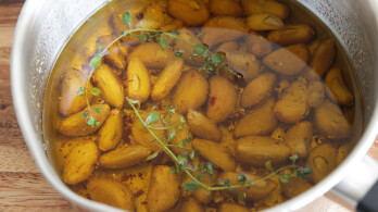 A pot filled with peeled garlic cloves submerged in olive oil, with sprigs of fresh thyme floating on top. The liquid is slightly golden, indicating the infusion of the garlic and herbs. The pot is placed on a wooden surface.