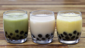 Three glasses of bubble tea are lined up on a wooden surface. The drinks include a green tea, a milk tea, and a mango or fruit tea, each with black tapioca pearls settled at the bottom.