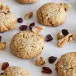 A batch of freshly baked cookies is spread out on a white surface. The cookies are surrounded by scattered pieces of walnuts and dried cranberries, suggesting they are ingredients in the cookies. The cookies appear golden brown and have a slightly cracked surface.