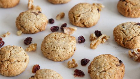 A batch of freshly baked cookies is spread out on a white surface. The cookies are surrounded by scattered pieces of walnuts and dried cranberries, suggesting they are ingredients in the cookies. The cookies appear golden brown and have a slightly cracked surface.