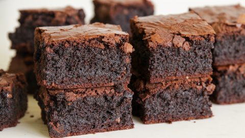 Close-up of several rich, chocolate brownies stacked on top of each other. The brownies have a moist and dense texture with a slightly flaky crust on top. The background is out of focus, drawing attention to the detailed texture of the baked goods.