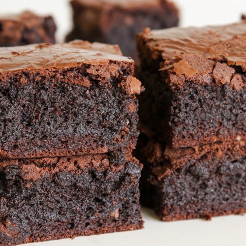 Close-up of several rich, chocolate brownies stacked on top of each other. The brownies have a moist and dense texture with a slightly flaky crust on top. The background is out of focus, drawing attention to the detailed texture of the baked goods.