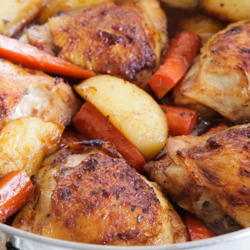 A close-up of a roasting pan filled with golden-brown, crispy chicken thighs, roasted potatoes, and carrots. The dish appears well-seasoned and freshly baked, with a slight sheen from the cooking juices.