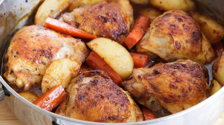 A close-up of a roasting pan filled with golden-brown, crispy chicken thighs, roasted potatoes, and carrots. The dish appears well-seasoned and freshly baked, with a slight sheen from the cooking juices.