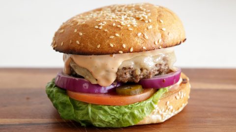 A close-up of a cheeseburger on a sesame seed bun. The burger is topped with melted cheese, red onion slices, pickles, creamy sauce, and layered over a slice of tomato and fresh green lettuce. The burger sits on a wooden surface.