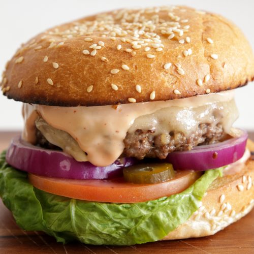 A close-up of a cheeseburger on a sesame seed bun. The burger is topped with melted cheese, red onion slices, pickles, creamy sauce, and layered over a slice of tomato and fresh green lettuce. The burger sits on a wooden surface.