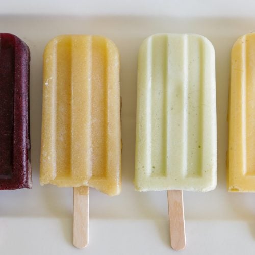 Four popsicles are lined up on a white surface. From left to right, they are deep red, light orange with a bite taken out, pale green, and creamy yellow in color. Each popsicle is on a wooden stick, and they all have a smooth, frozen texture.