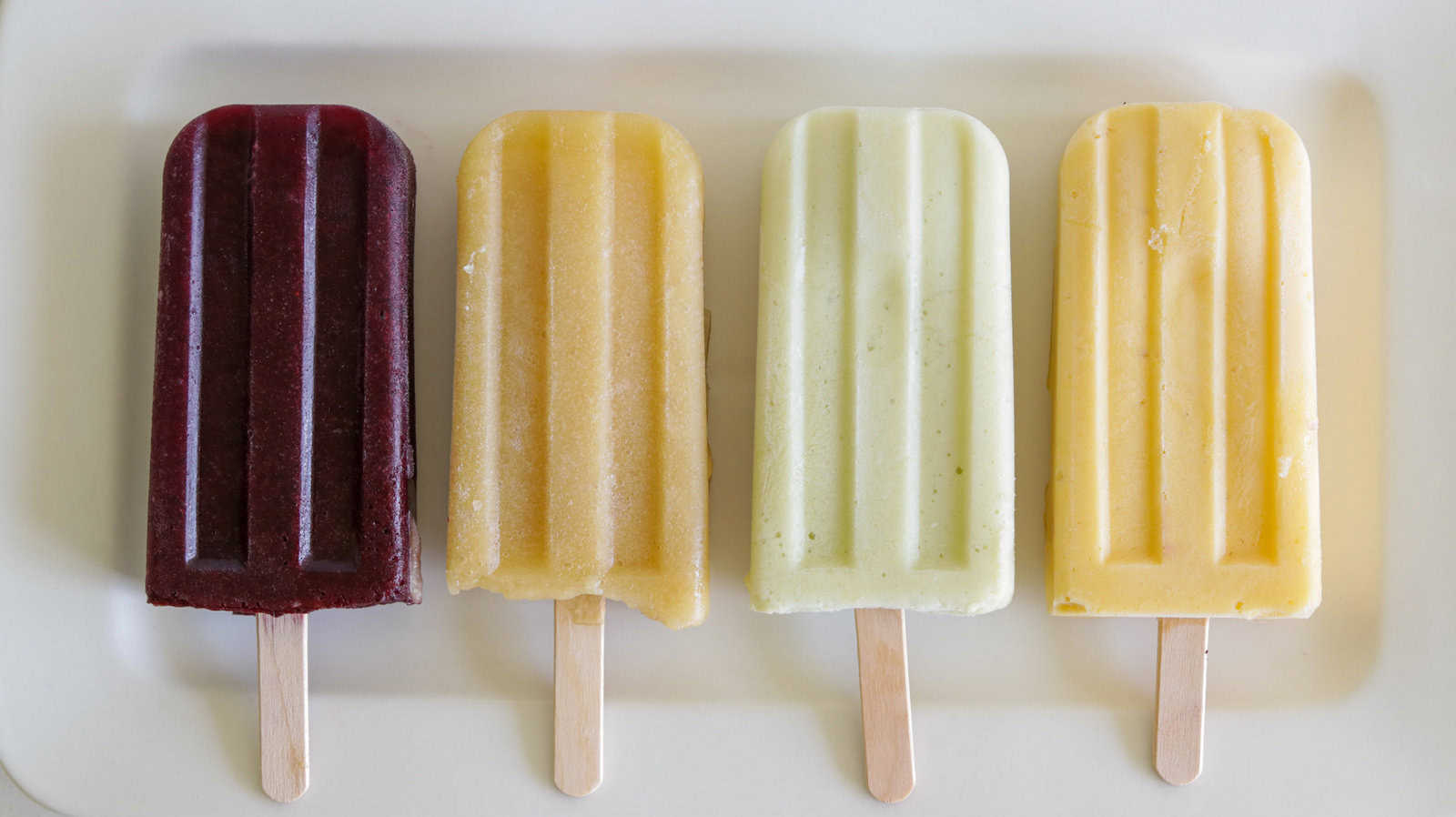 Four popsicles are lined up on a white surface. From left to right, they are deep red, light orange with a bite taken out, pale green, and creamy yellow in color. Each popsicle is on a wooden stick, and they all have a smooth, frozen texture.