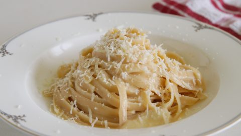 A plate of creamy fettuccine Alfredo topped with shredded Parmesan cheese. The dish is served on a decorative white plate, with a red and white checkered cloth napkin in the background. The pasta looks rich and indulgent, glistening with sauce.