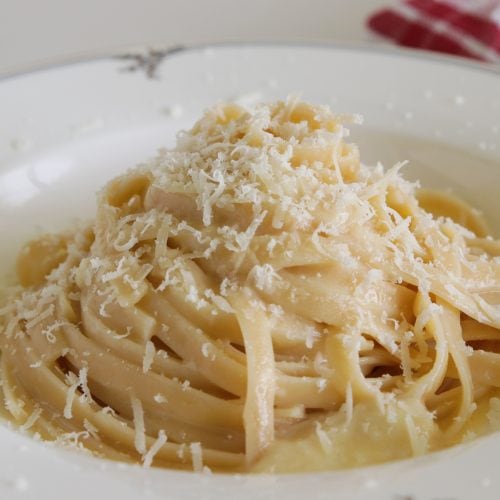 A plate of creamy fettuccine Alfredo topped with shredded Parmesan cheese. The dish is served on a decorative white plate, with a red and white checkered cloth napkin in the background. The pasta looks rich and indulgent, glistening with sauce.