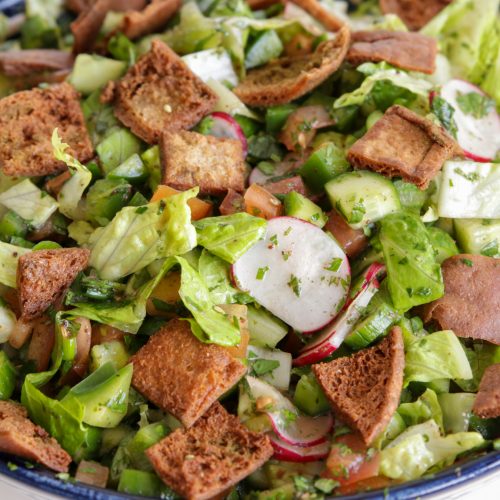 A bowl of fresh fattoush salad showcasing chopped lettuce, cucumbers, radishes, tomatoes, bell peppers, and parsley, topped with crispy toasted pita bread pieces. The vegetables are finely diced and mixed together, highlighting a colorful and vibrant presentation.