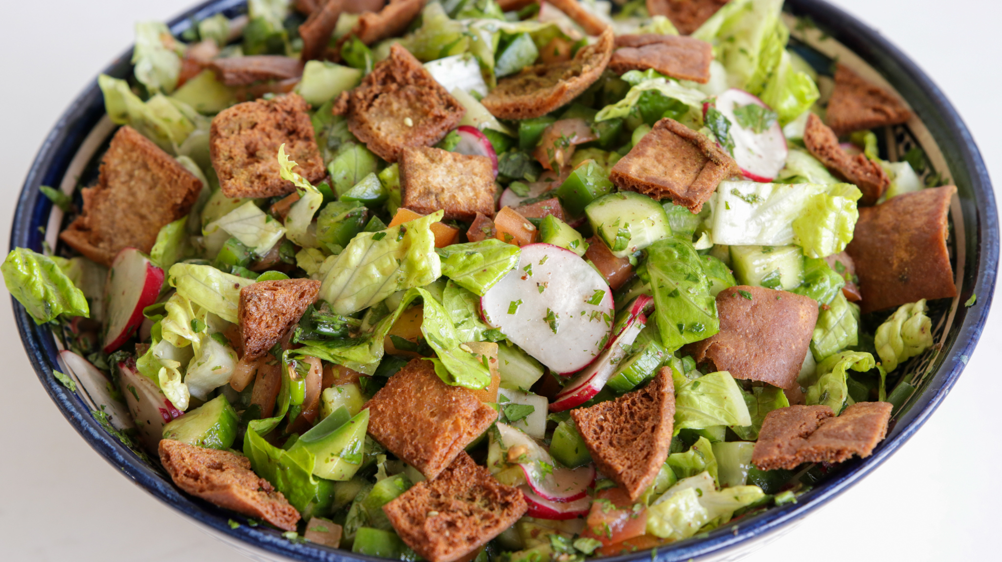 A bowl of fresh fattoush salad showcasing chopped lettuce, cucumbers, radishes, tomatoes, bell peppers, and parsley, topped with crispy toasted pita bread pieces. The vegetables are finely diced and mixed together, highlighting a colorful and vibrant presentation.