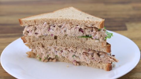 A sandwich made with two triangular slices of whole wheat bread, filled with a mixture of tuna, chopped red onions, and lettuce, is placed on a white plate. The sandwich is cut into halves and stacked on top of each other on a wooden table background.